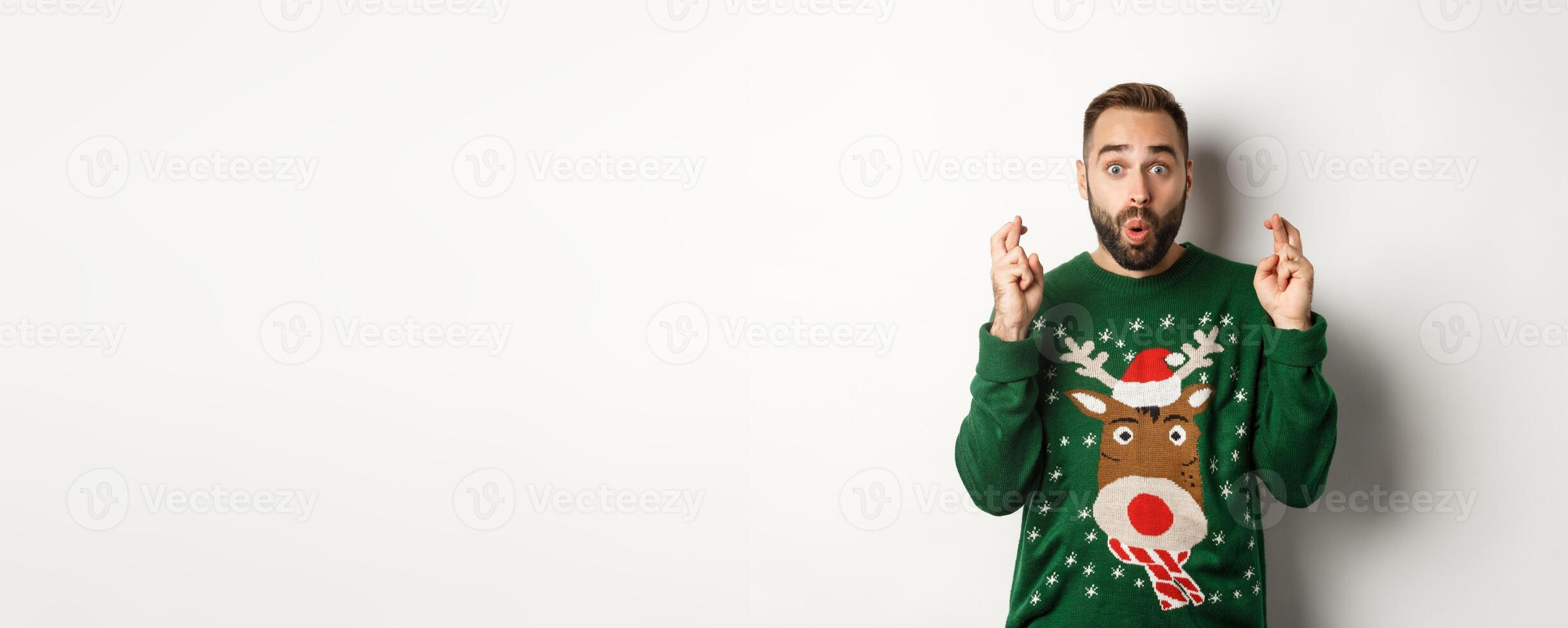 Christmas, holidays and celebration. Excited guy making wish, keep fingers crossed for good luck and smiling, standing over white background photo