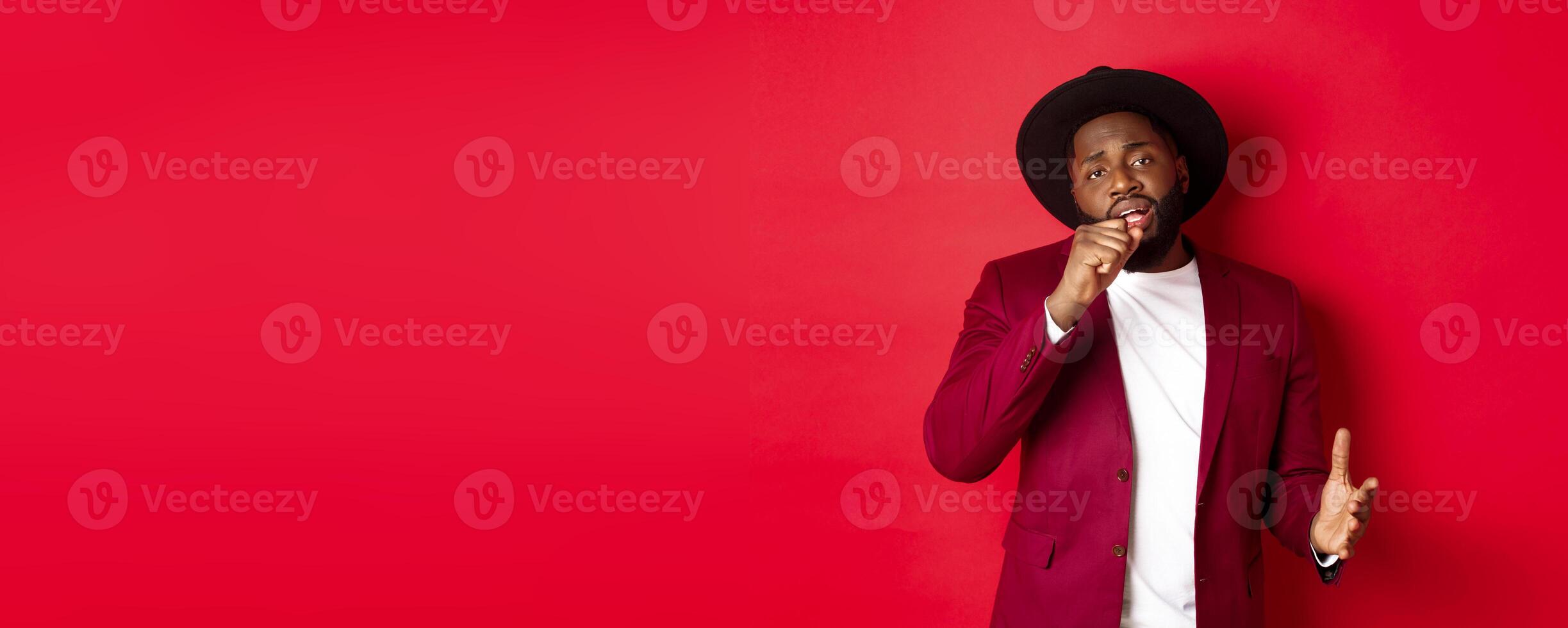 Christmas shopping and people concept. Handsome Black male model in party outfit singing in invisible microphone, looking passionate at camera, red background photo