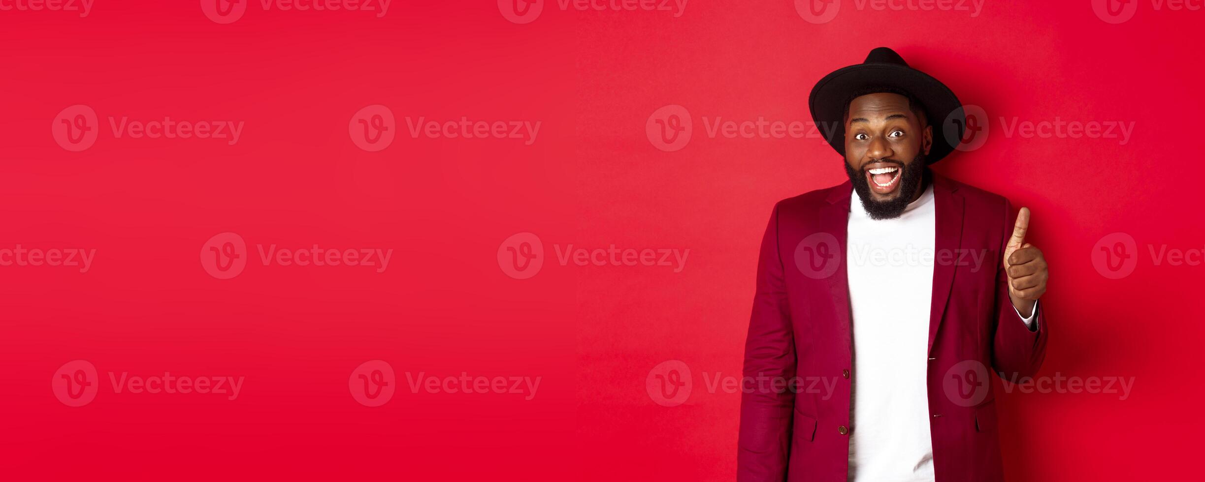 Christmas shopping and people concept. Handsome bearded Black man in party blazer showing thumb up, looking amazed and satisfied, red background photo