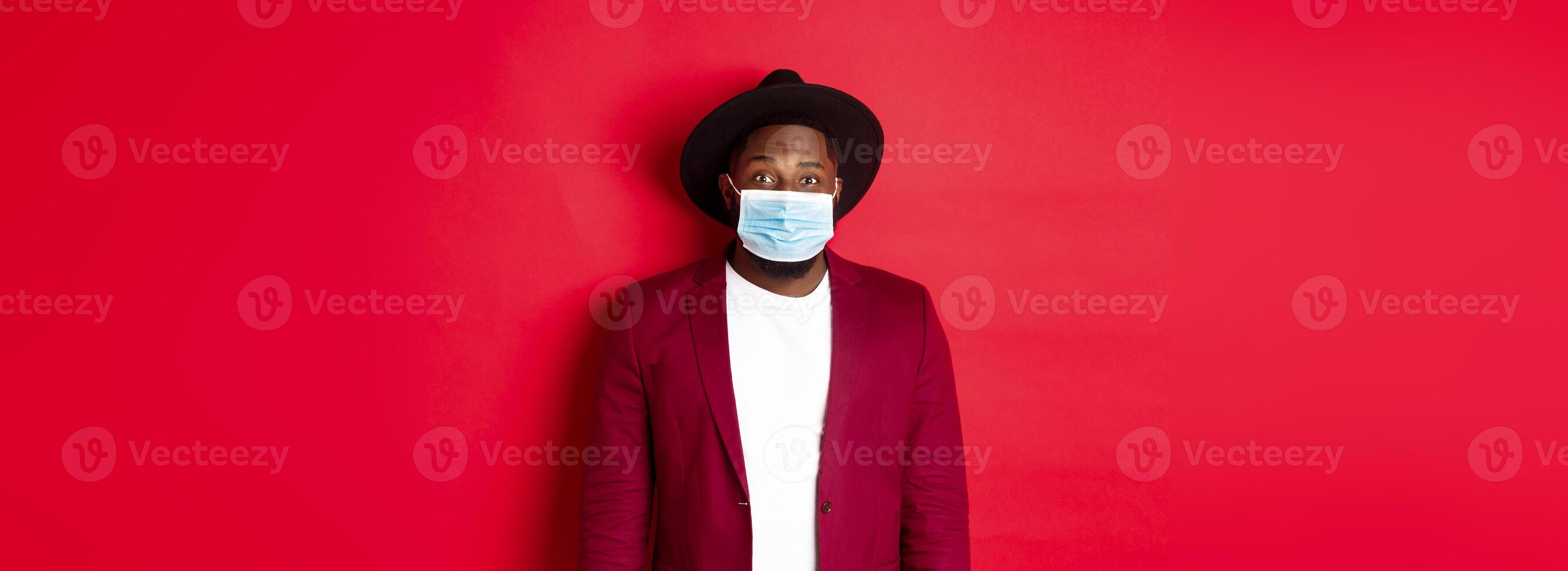 Covid-19 and fashion concept. Handsome african american man in stylish blazer and hat, wearing medical mask from coronavirus, standing over red background photo