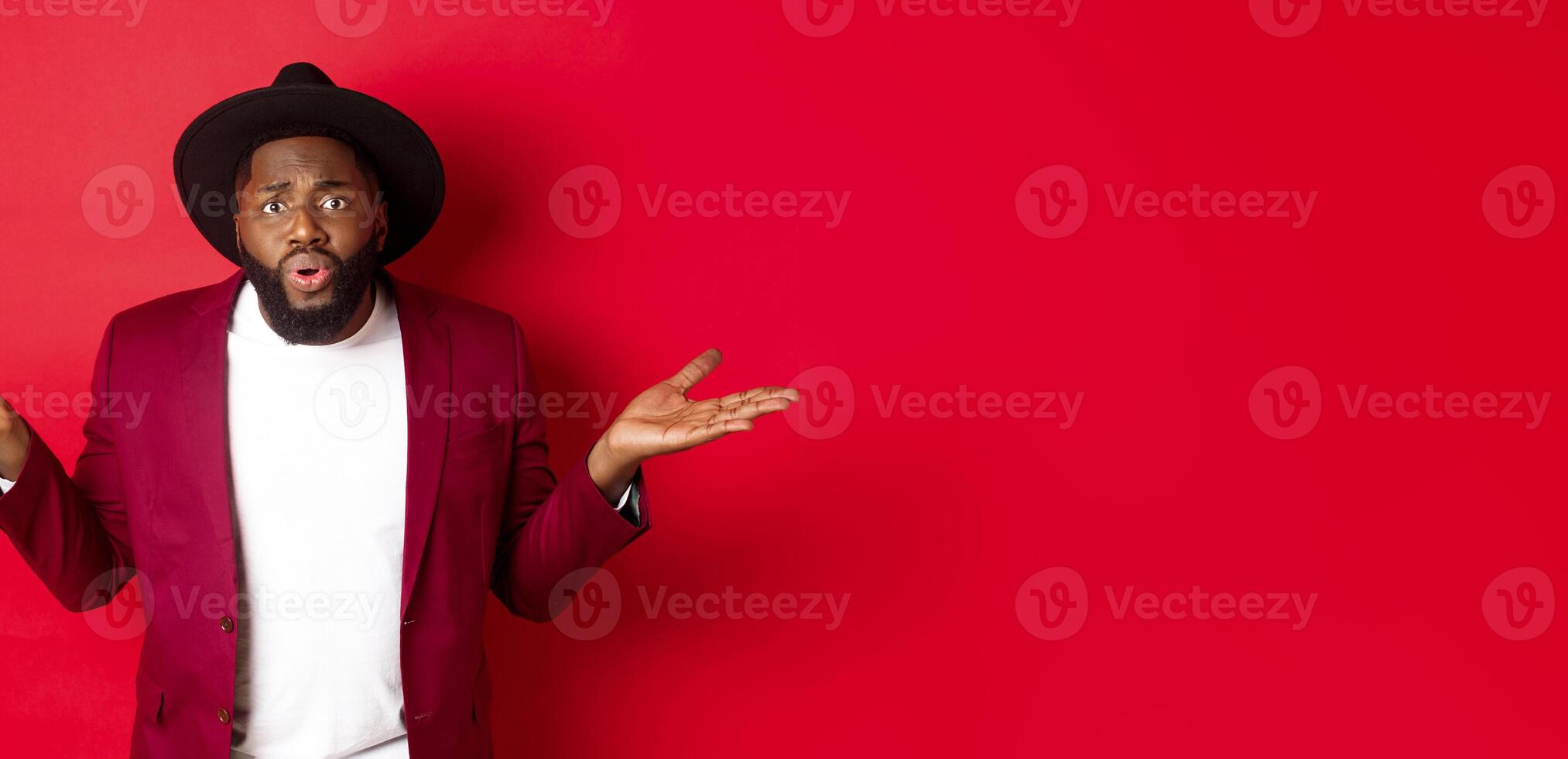 Confused and worried Black man spread hands sideways, staring at camera puzzled, asking what wrong, standing against red background photo