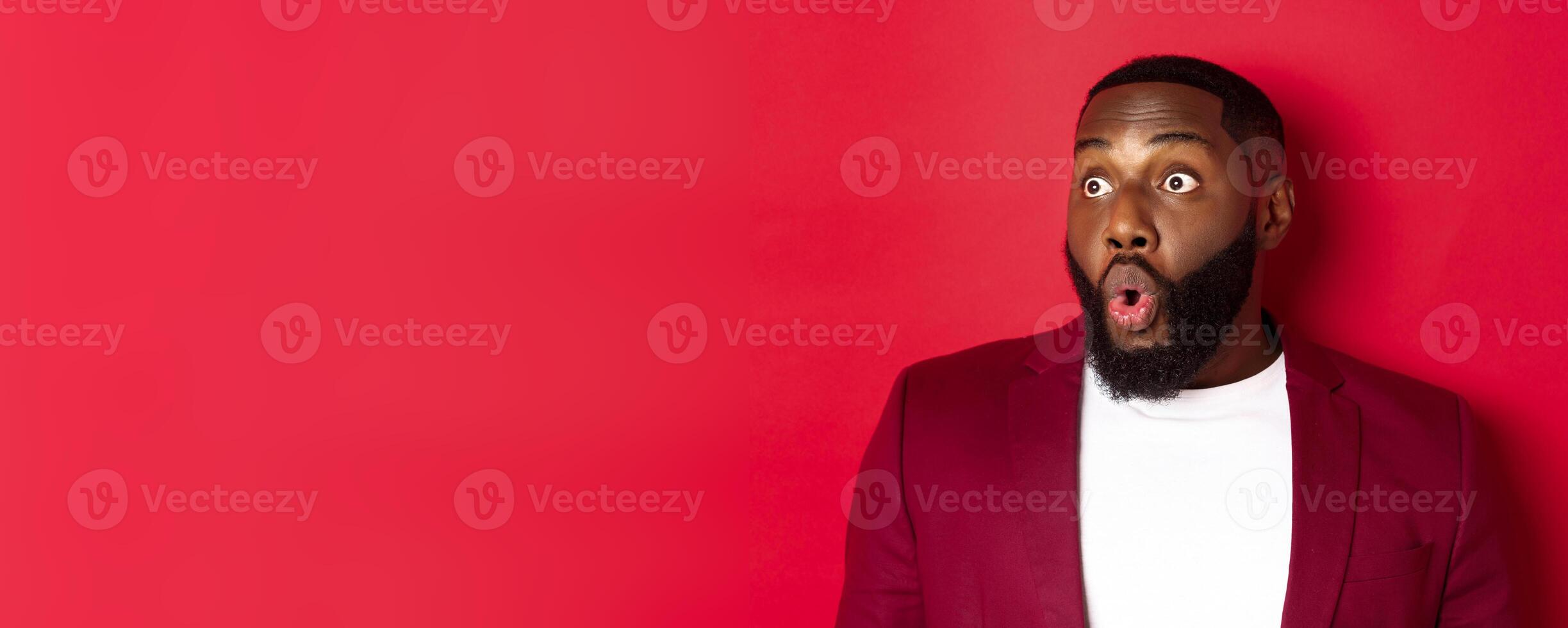 Close-up of impressed Black man looking left at copy space, saying wow and staring with amazement, standing over red background photo