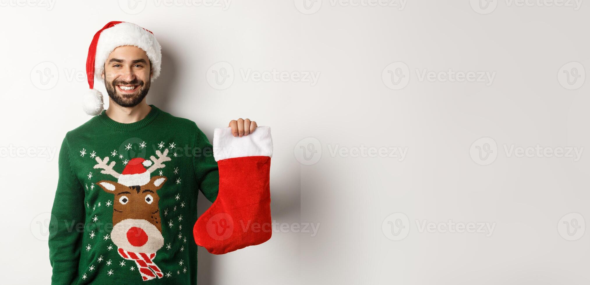 Xmas party and holidays concept. Happy man in Santa hat bringing gifts in Christmas sock and smiling, standing over white background photo