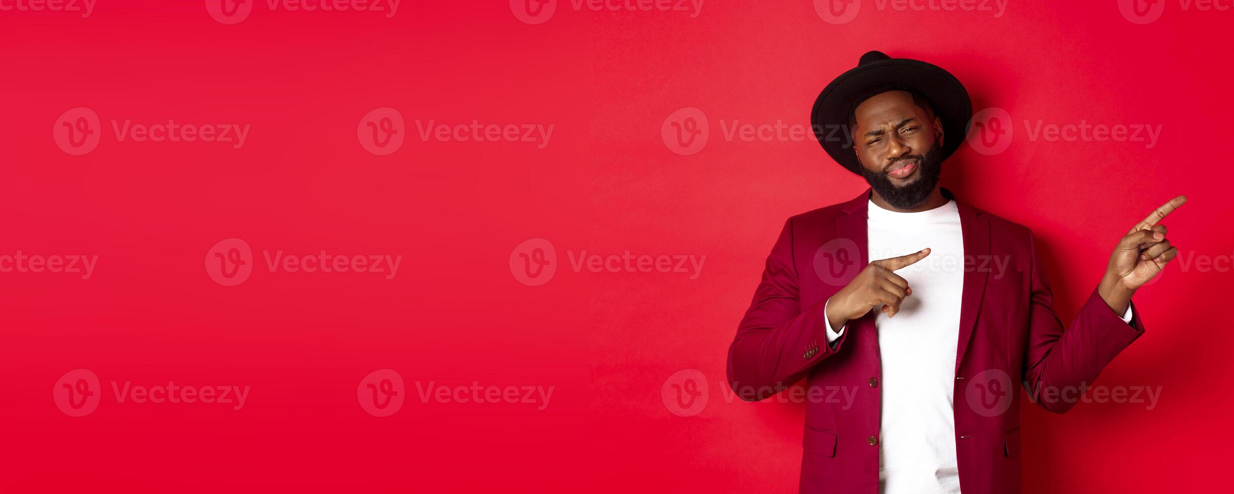 Winter holidays and shopping concept. Skeptical and unamused Black man pointing fingers right and grimacing displeased, do not recommend offer, standing over red background photo