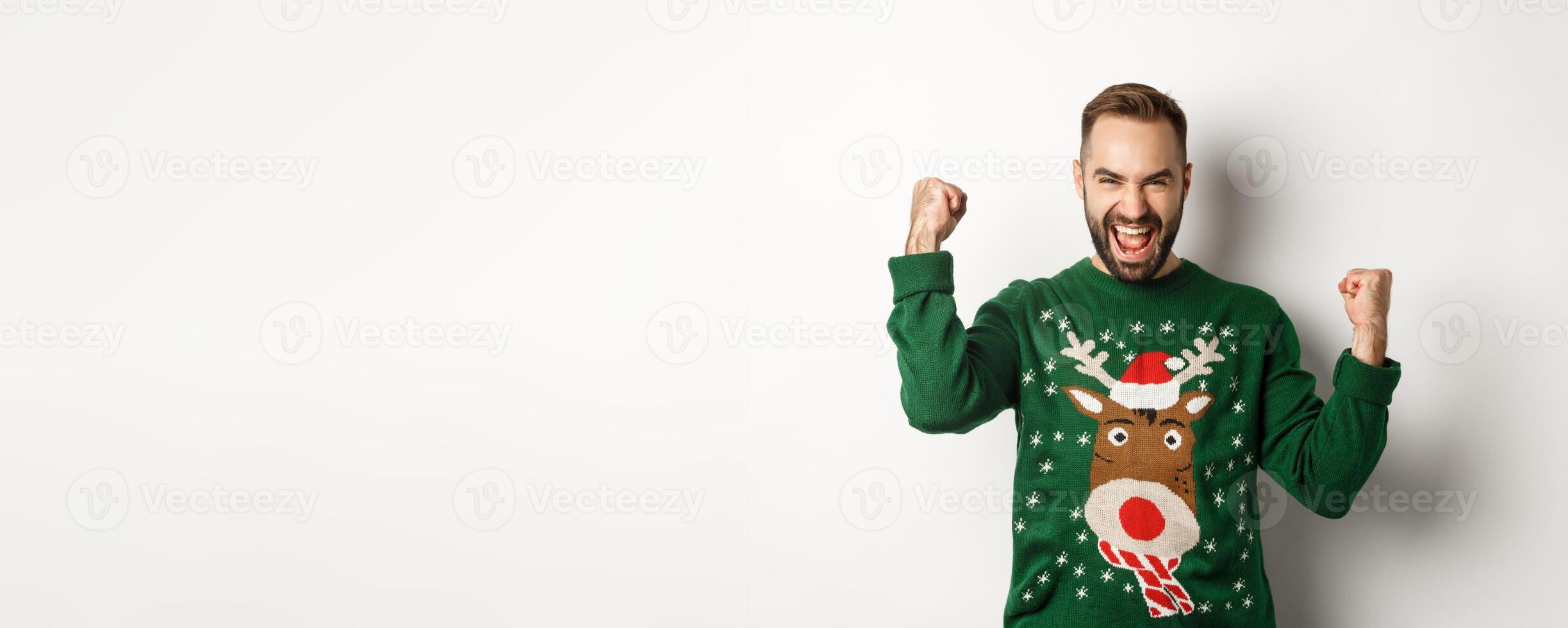 New Year, holidays and celebration. Excited bearded man in Christmas sweater, making fist pumps and shouting for joy, rejoicing and triumphing, white background photo