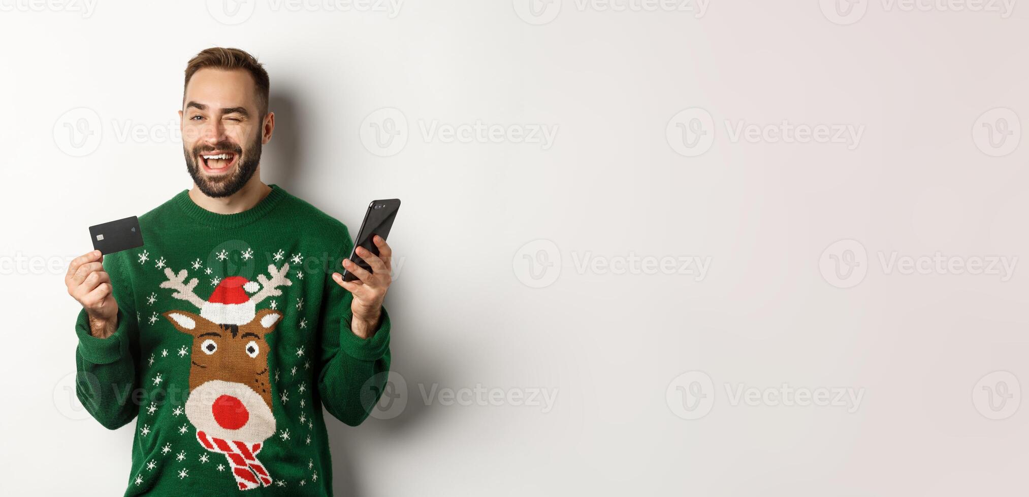 New Year, online shopping and christmas concept. Happy bearded man using mobile phone and credit card, winking at camera, order something in internet, white background photo