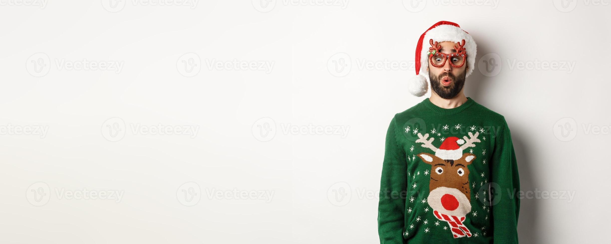 Christmas holidays, celebration concept. Surprised guy in party glasses and santa hat looking down amused, standing over white background photo
