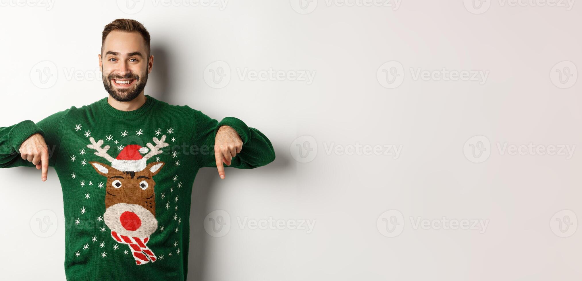 Winter holidays and christmas. Handsome smiling man with beard, pointing fingers down and showing promo offer, wearing green sweater, standing over white background photo