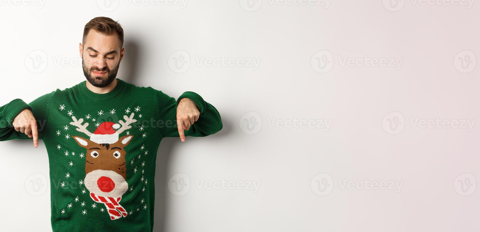 Winter holidays and christmas. Skeptical bearded man in green sweater, pointing and looking down displeased, standing over white background photo