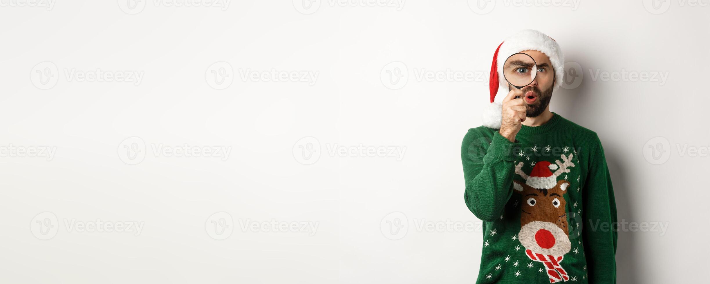 Christmas and holidays concept. Curious guy looking at something through magnifying glass, standing in Santa hat with xmas sweater, standing over white background photo