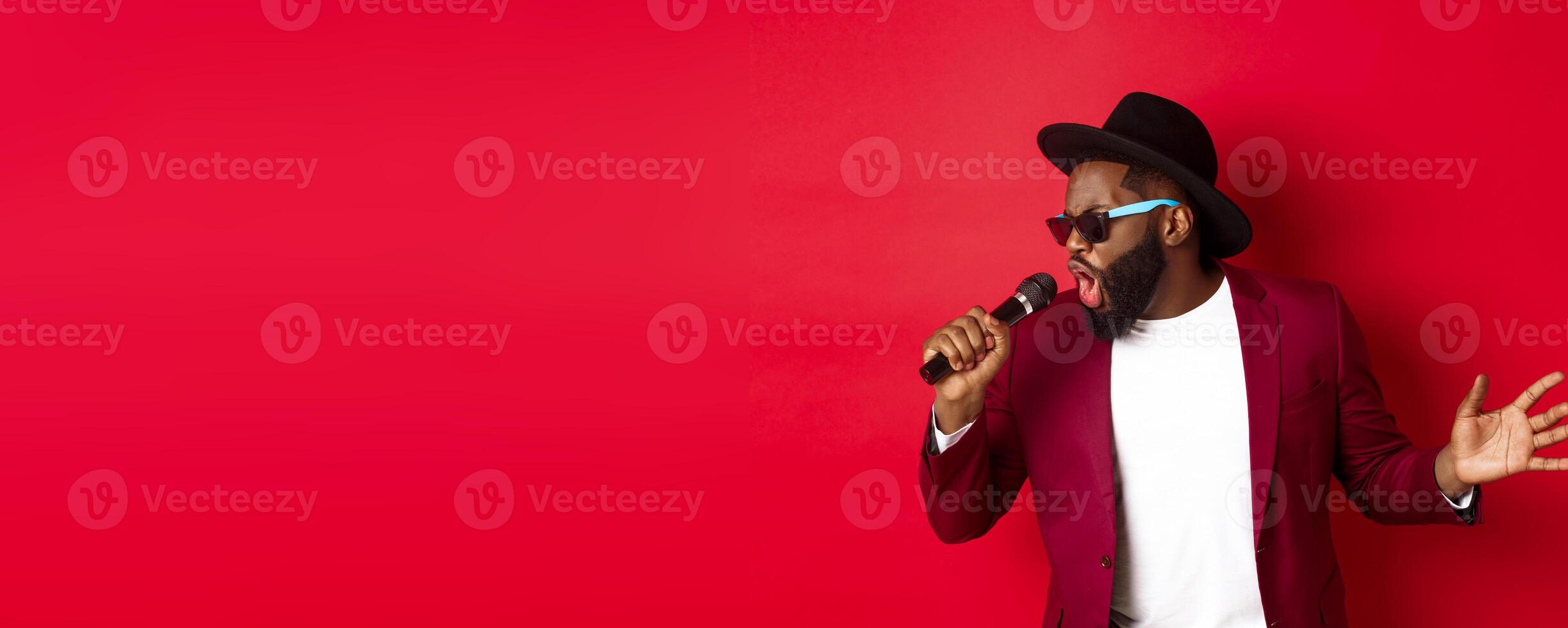 Passionate black male singer performing against red background, singing into microphone, wearing party outfit, standing over red background photo