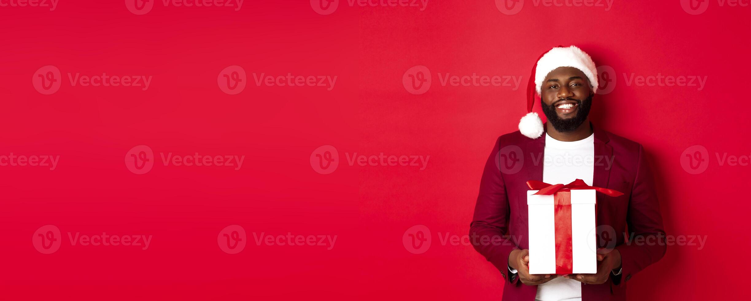 Christmas, New Year and shopping concept. Happy bearded african american man holding xmas present, smiling at camera, standing in santa hat against red background photo