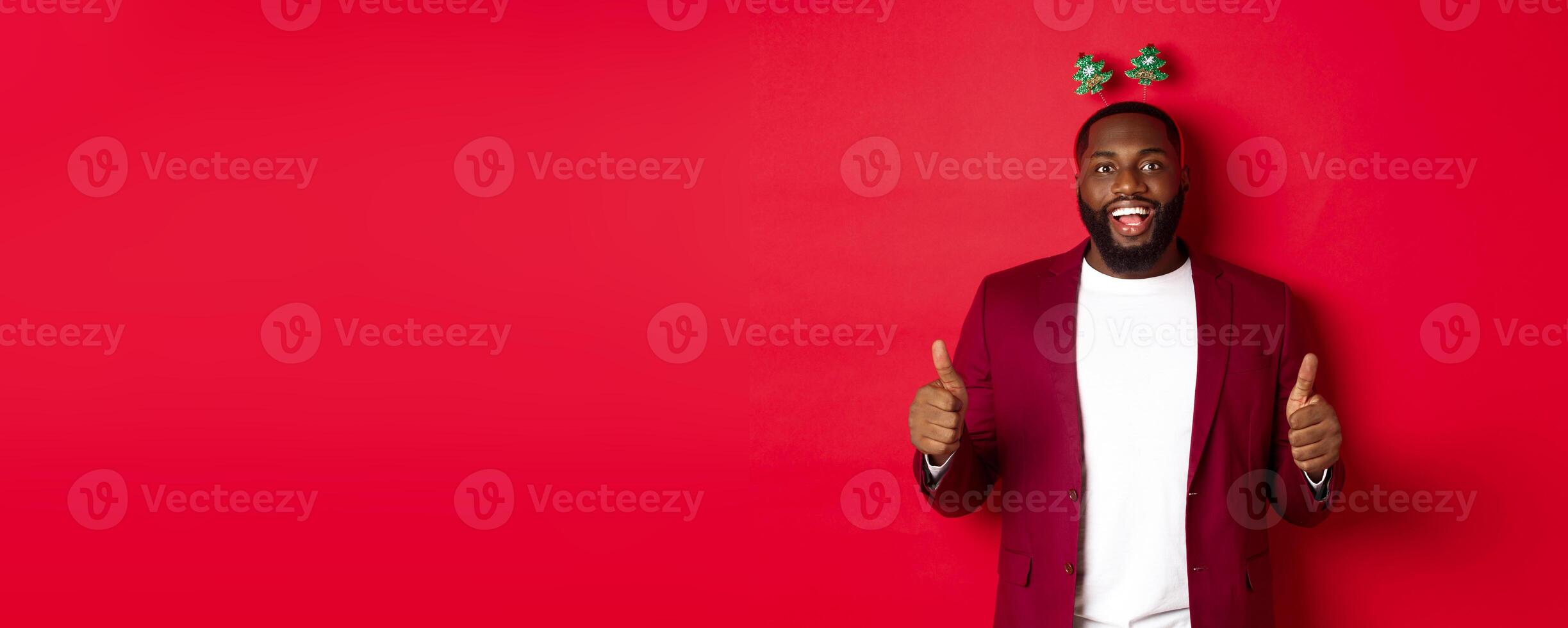 Merry Christmas. Happy african american man celebarting New Year, wearing funny party headband and showing thumb up, like and praise something, standing over red backgrond photo