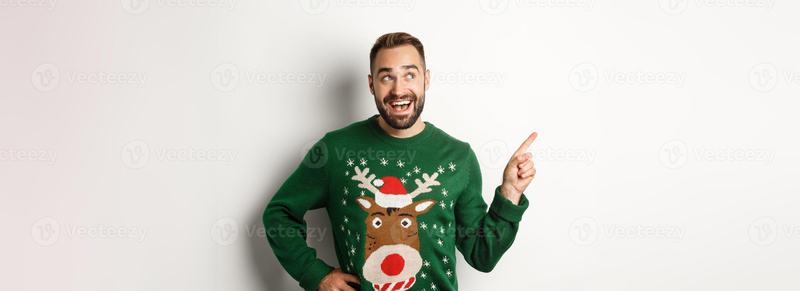 New year celebration and winter holidays concept. Dreamy bearded man in green christmas sweater, pointing at upper left corner and smiling amused, white background photo