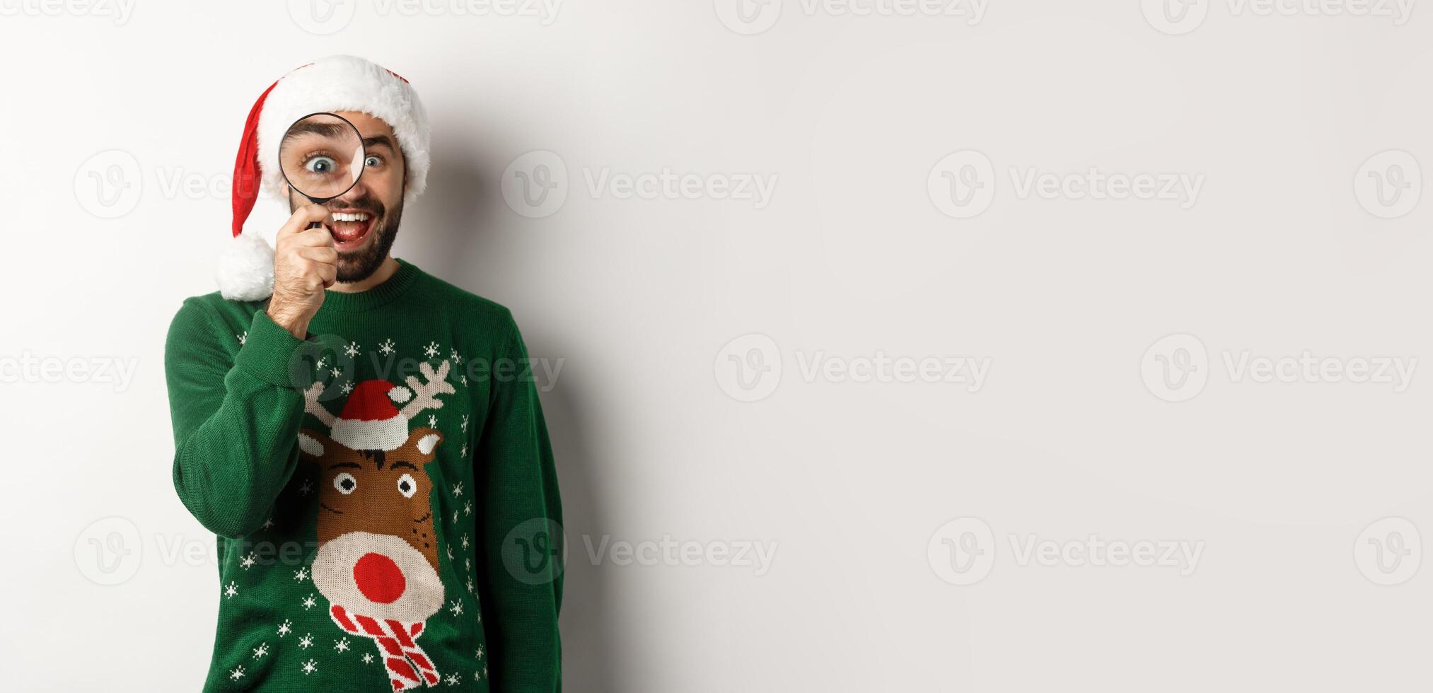 Christmas and holidays concept. Funny bearded guy in Santa hat looking through magnifying glass with amazement, found something, standing over white background photo