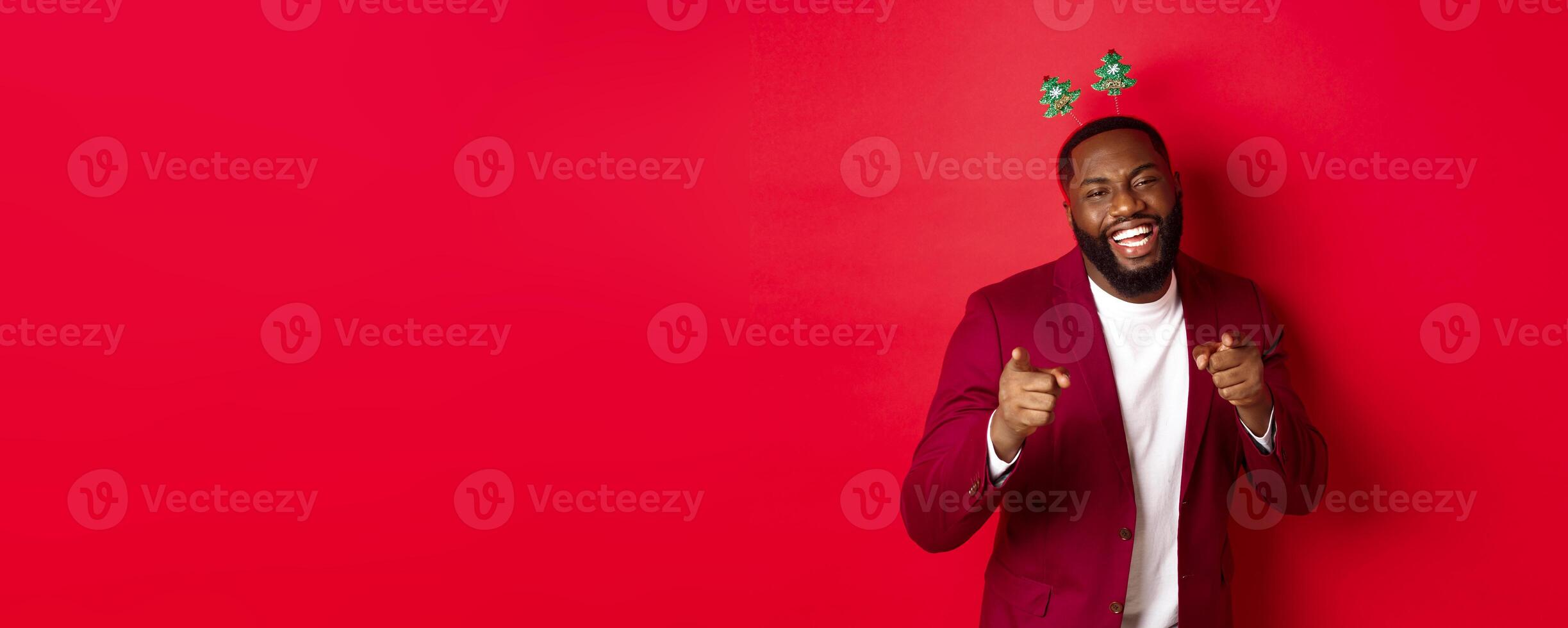 Merry Christmas. Handsome african american man in blazer and party headband, celebrating new year, pointing at camera and smiling, congratulating, red background photo