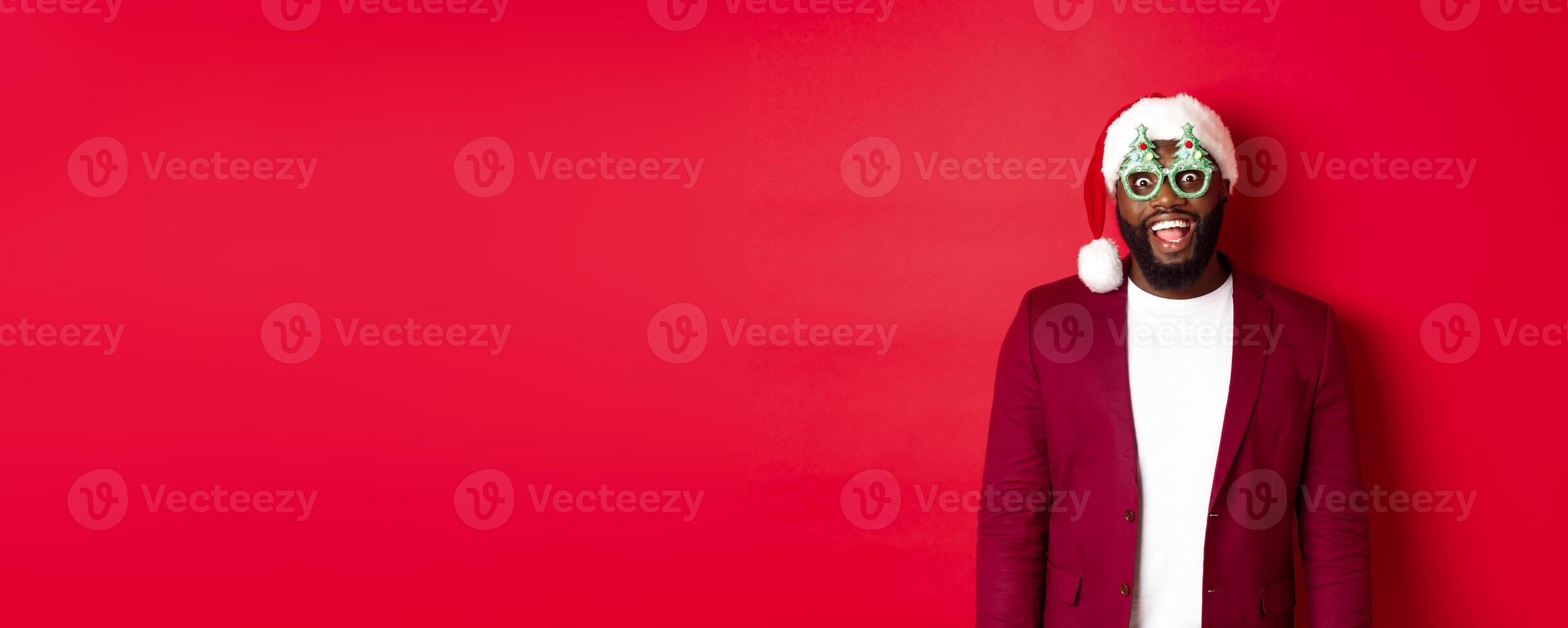 Merry Christmas. Cheerful Black man wearing funny party glasses and santa hat, smiling joyful, celebrating winter holidays, standing over red background photo