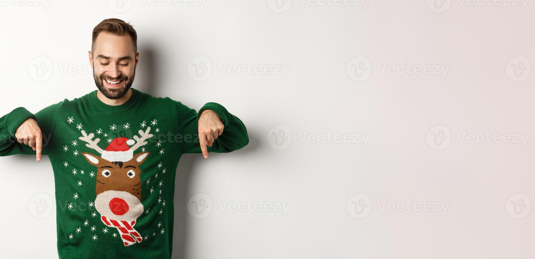 Winter holidays and christmas. Happy caucasian man in green sweater, pointing fingers down and looking satisfied, standing over white background photo