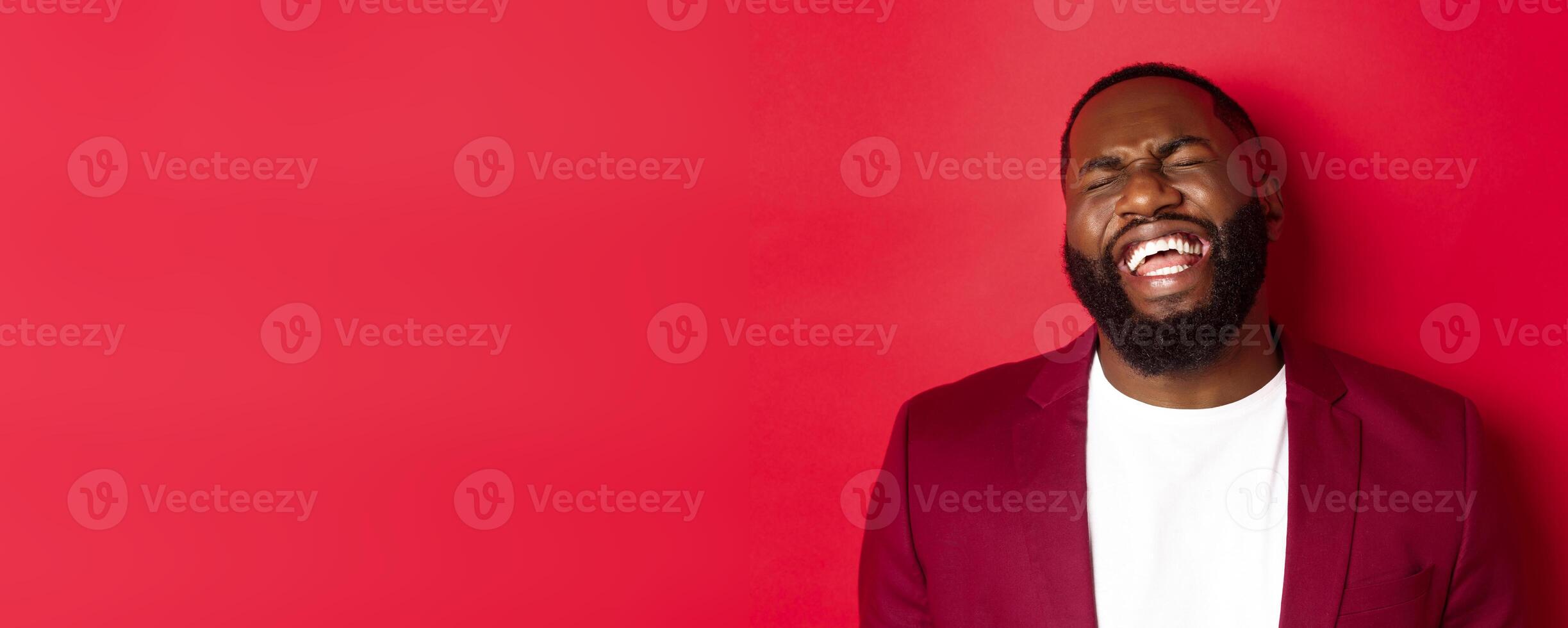 Close-up of happy and carefree Black man having fun, laughing and smiling, standing in blazer against red background photo