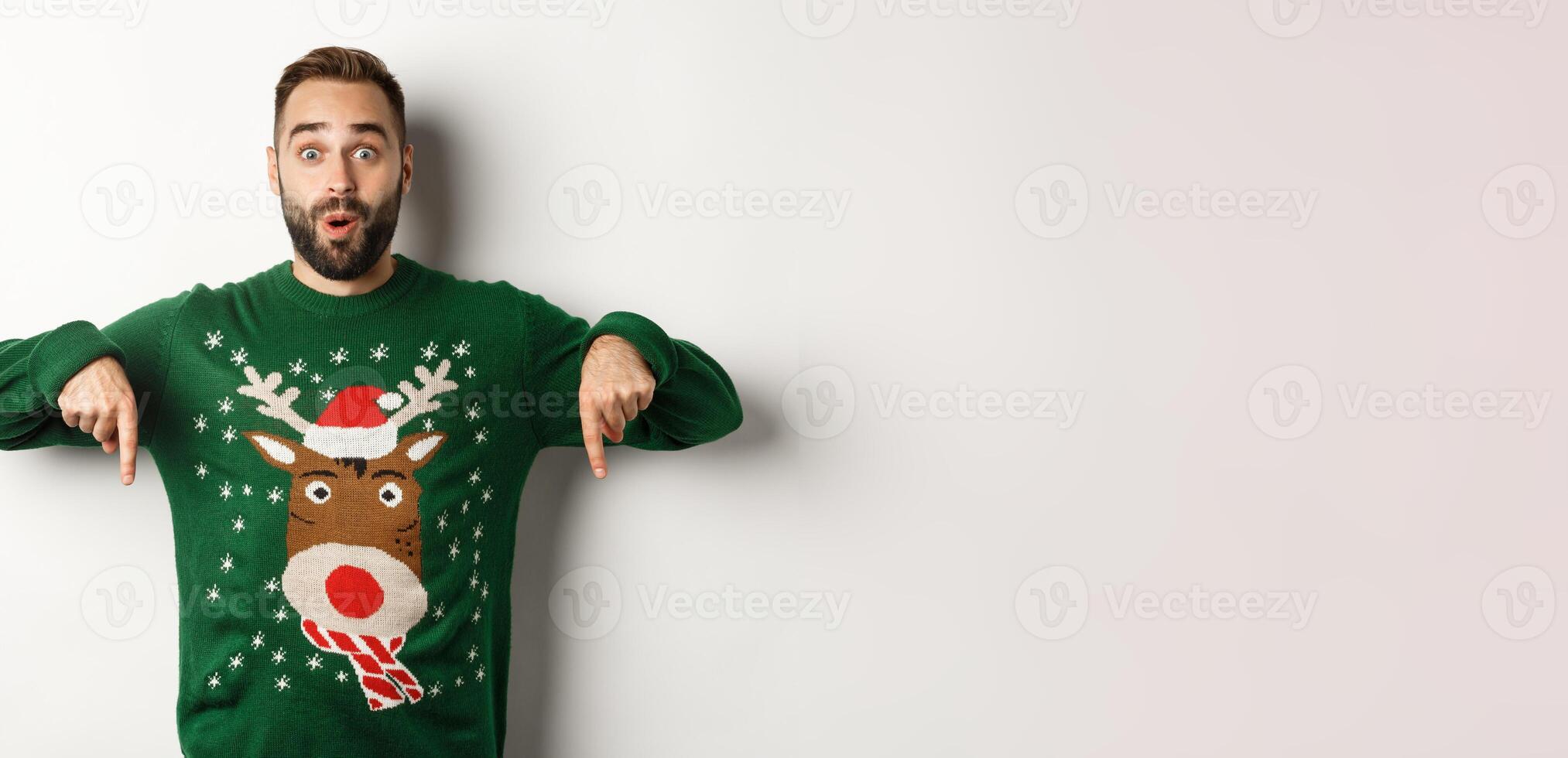 Winter holidays and christmas. Surprised bearded man in green sweater showing announcement, pointing fingers down at promo banner, white background photo