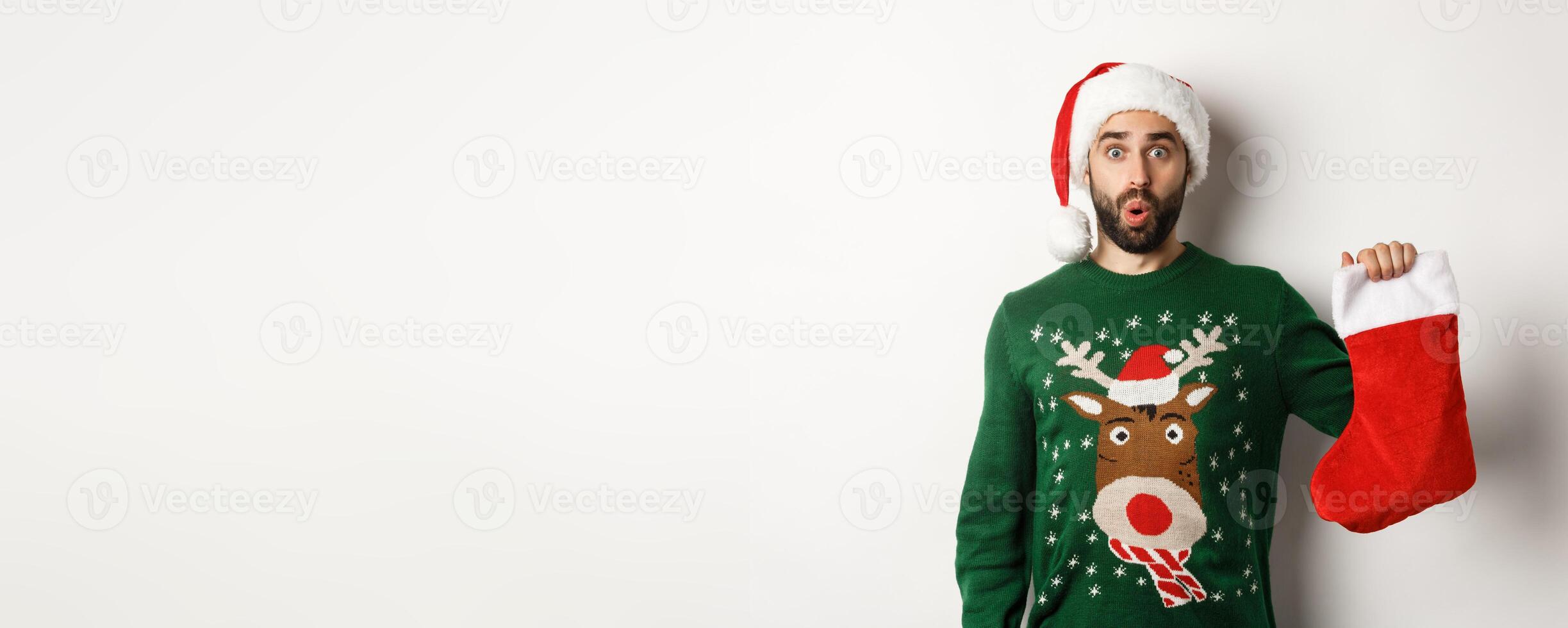 Xmas and winter holidays concept. Happy man got a gift in Christmas sock, looking excited, standing in Santa hat against white background photo