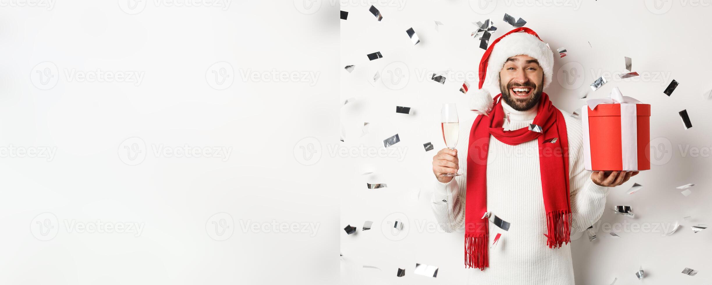 Christmas party and holidays concept. Happy guy celebrating New Year, holding gift and glass, drinking champagne and laughing, confetti flying around, white background photo