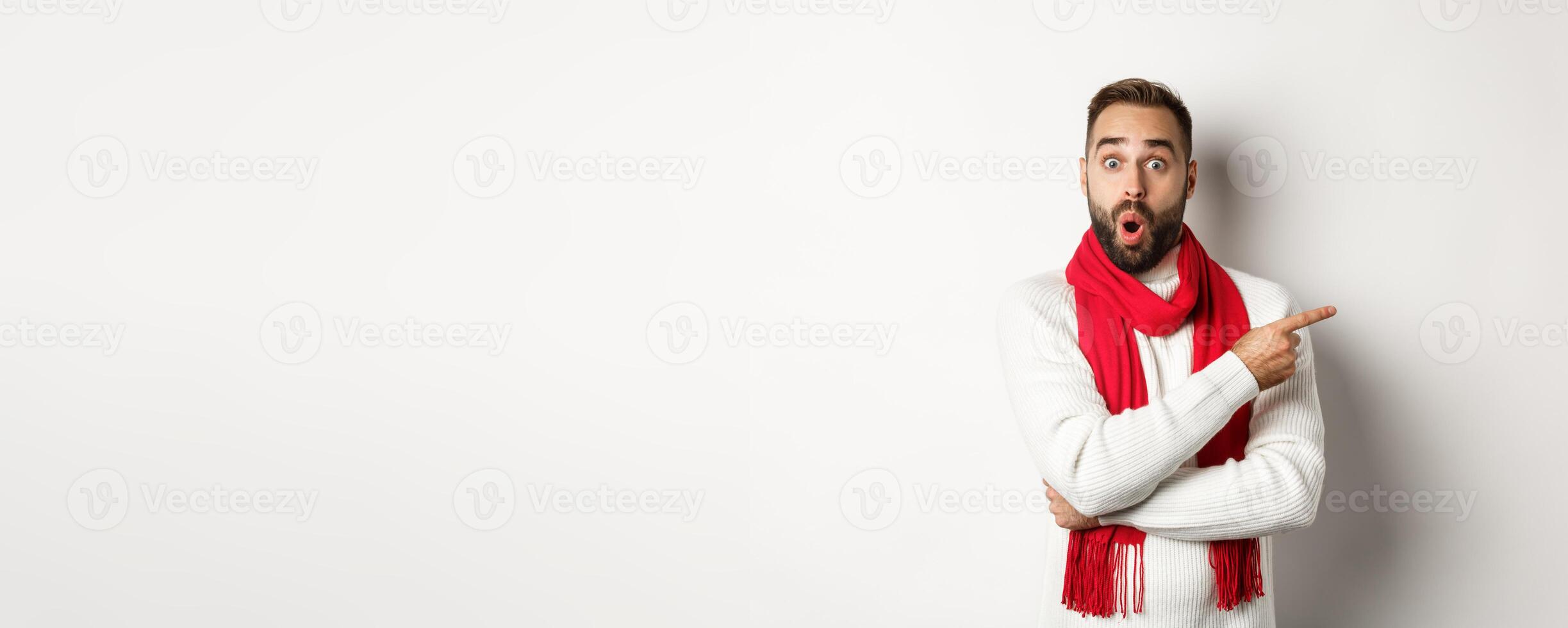 Christmas holidays and celebration concept. Surprised bearded man pointing finger right and looking impressed at camera, standing in red scarf and sweater, white background photo