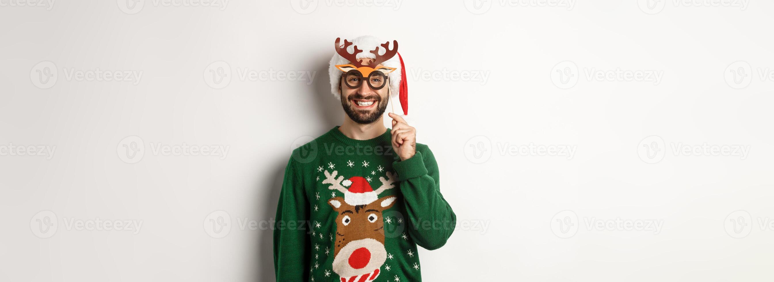 Christmas and holidays concept. Smiling beared man in Santa hat looking happy, holding party mask for New Year, celebrating xmas, standing over white background photo