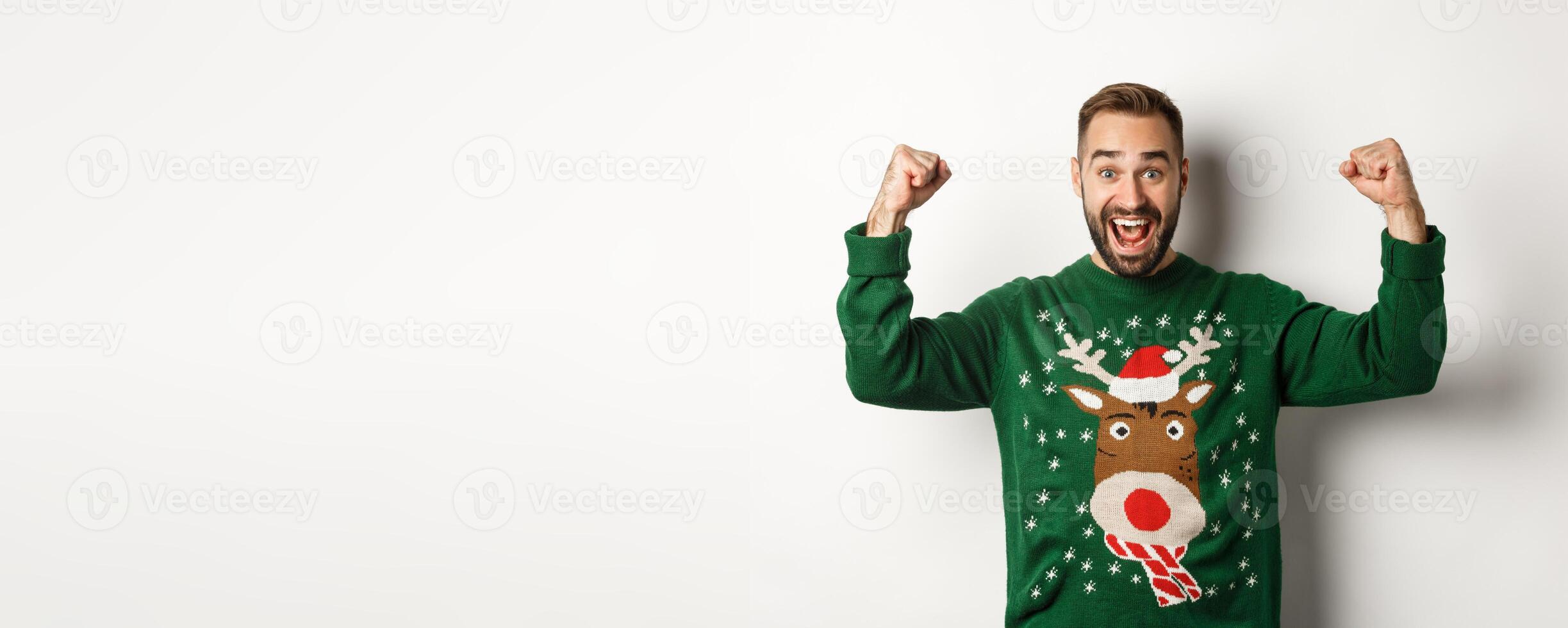 New Year, holidays and celebration. Relieved and excited man in Christmas sweater, raising hands up and celebrating, triumphing, standing over white background photo