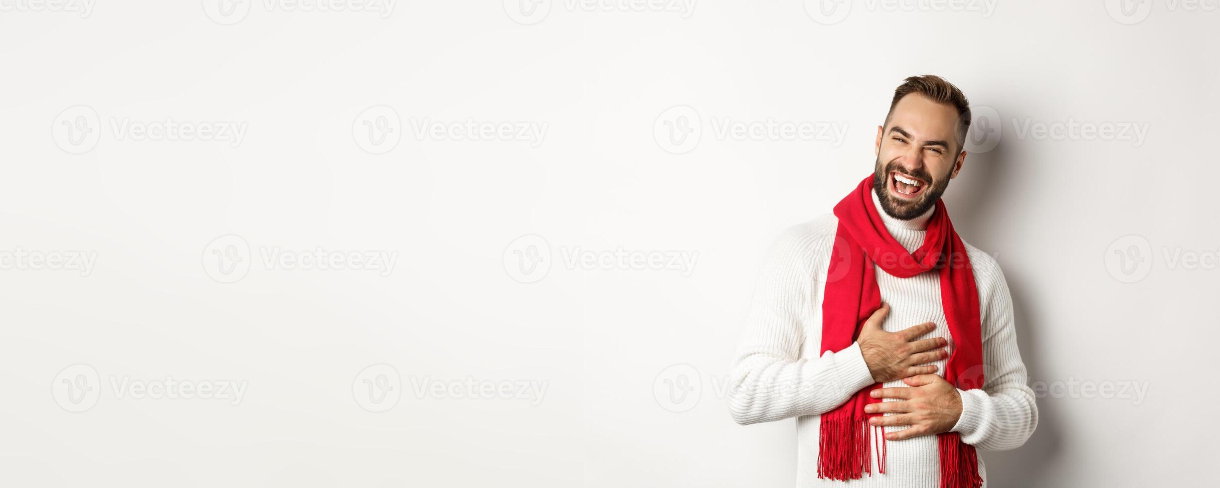 Happy man laughing and touching belly, chuckling from funny joke, standing in winter sweater and red scarf, white background photo