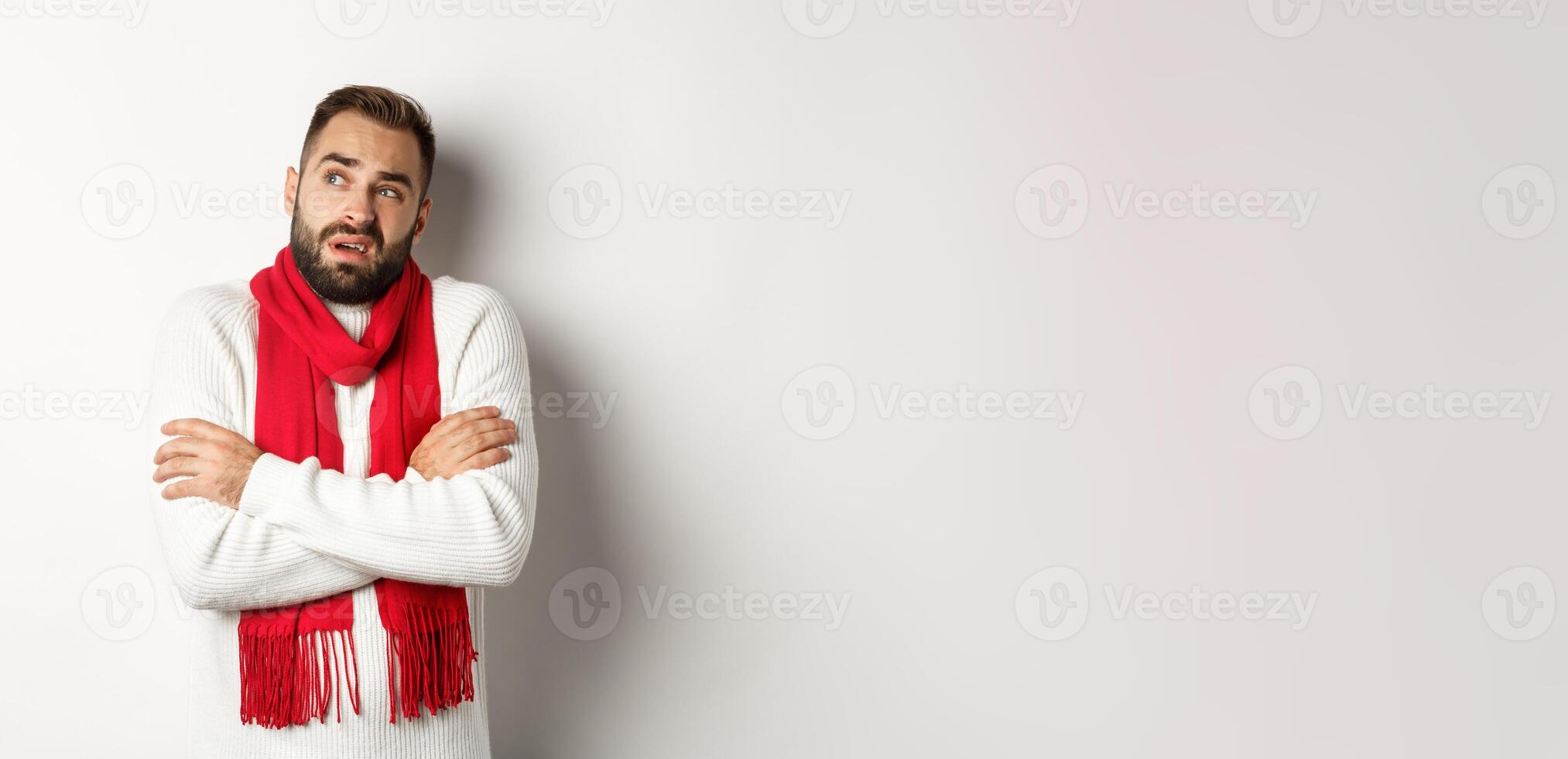 Confused man looking doubtful, standing with hands crossed and feeling cold on winter, wearing christmas sweater with red scarf, standing over white background photo