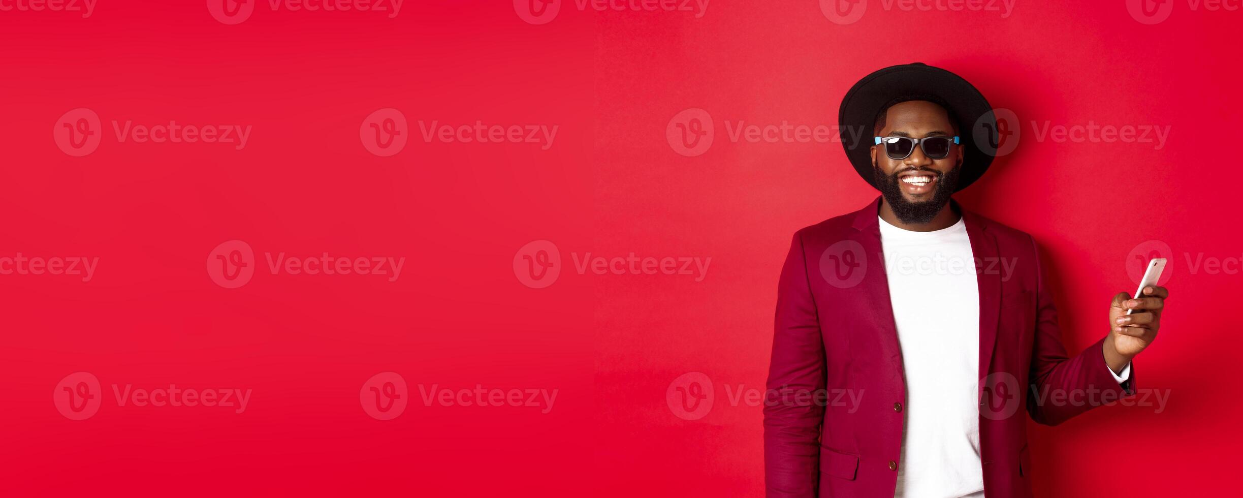 Handsome african american man shopping online, making an order on smartphone and smiling, standing over red background photo