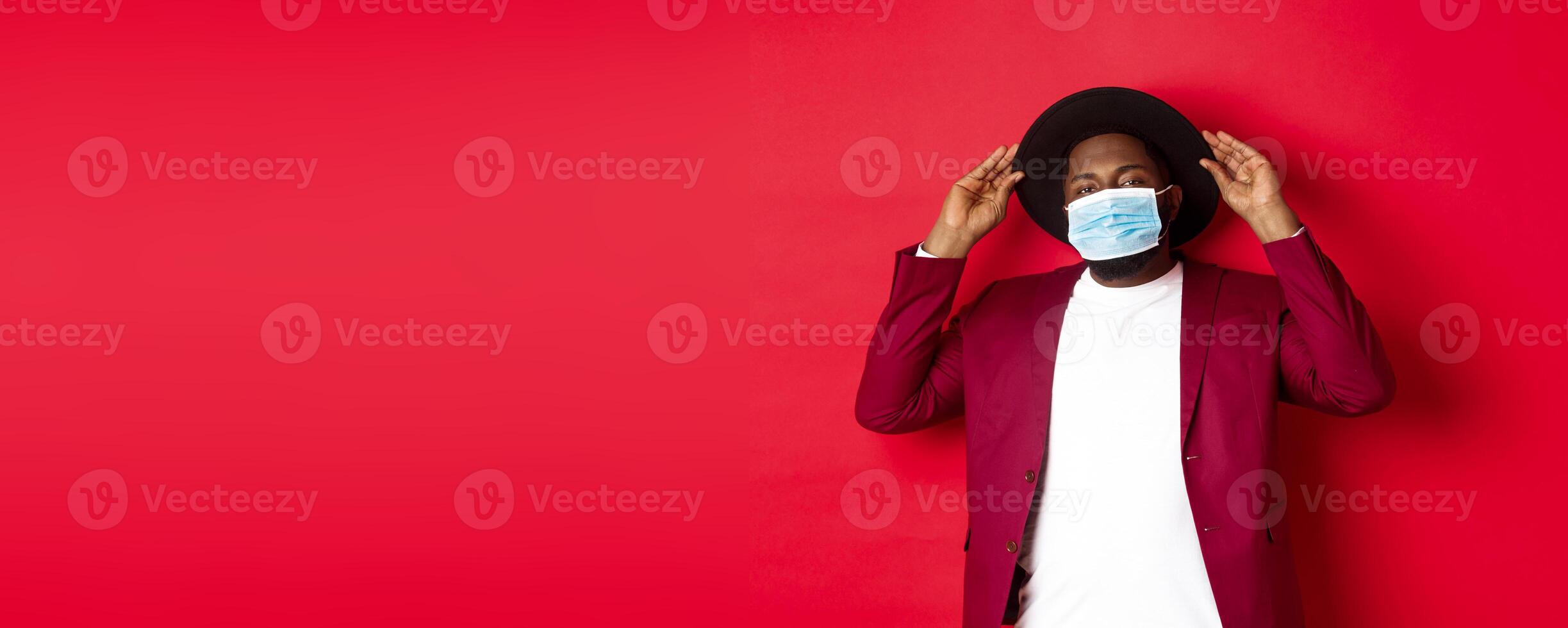Covid-19, quarantine and holidays concept. Handsome and stylish african american man in face mask, put hat on head and looking sassy, dress-up for party, standing over red background photo