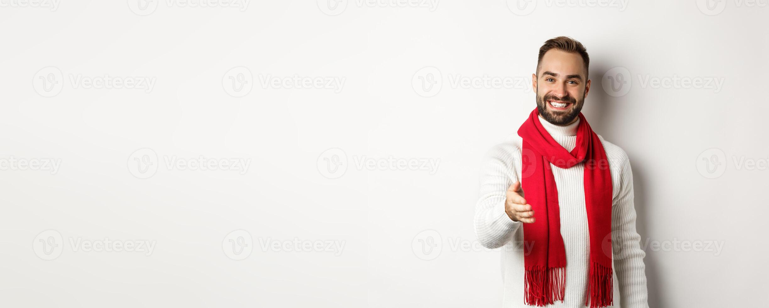 Friendly young man greeting person with handshake, smiling and wishing merry christmas, wearing red scarf and winter sweater, white background photo