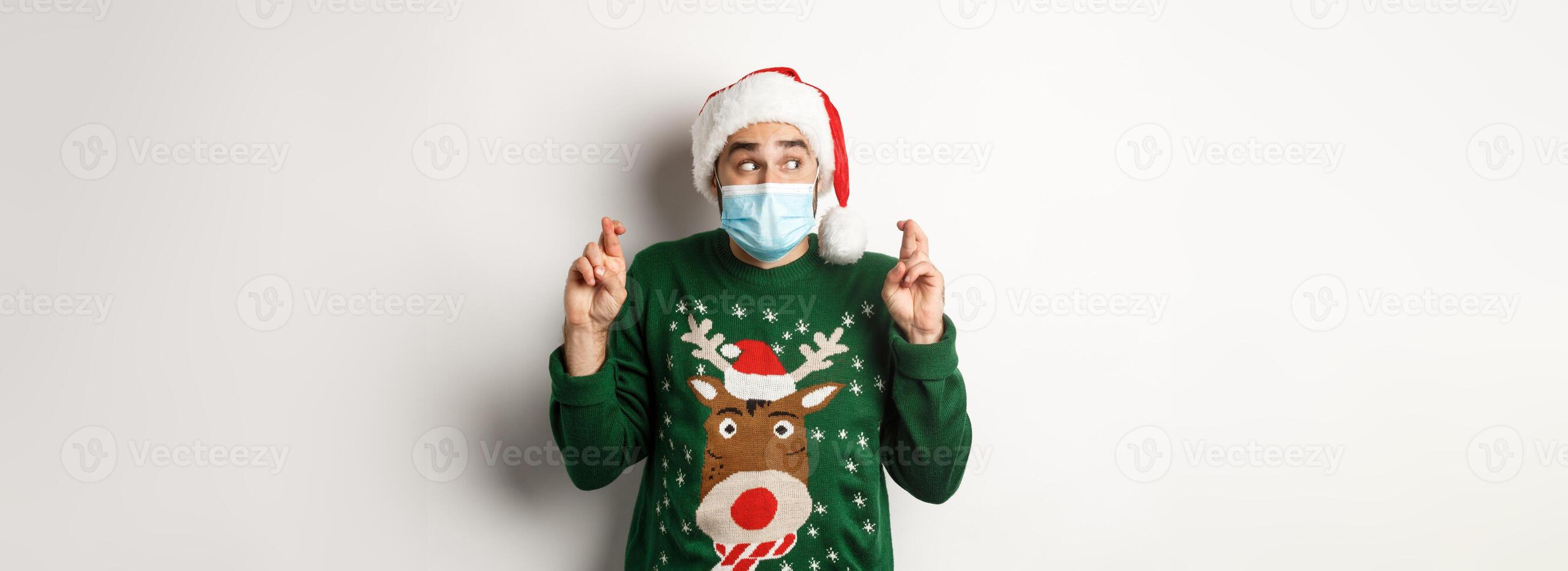 Concept of covid-19 and Christmas holidays. Excited man in face mask and Santa hat cross fingers, making a wish, standing over white background photo