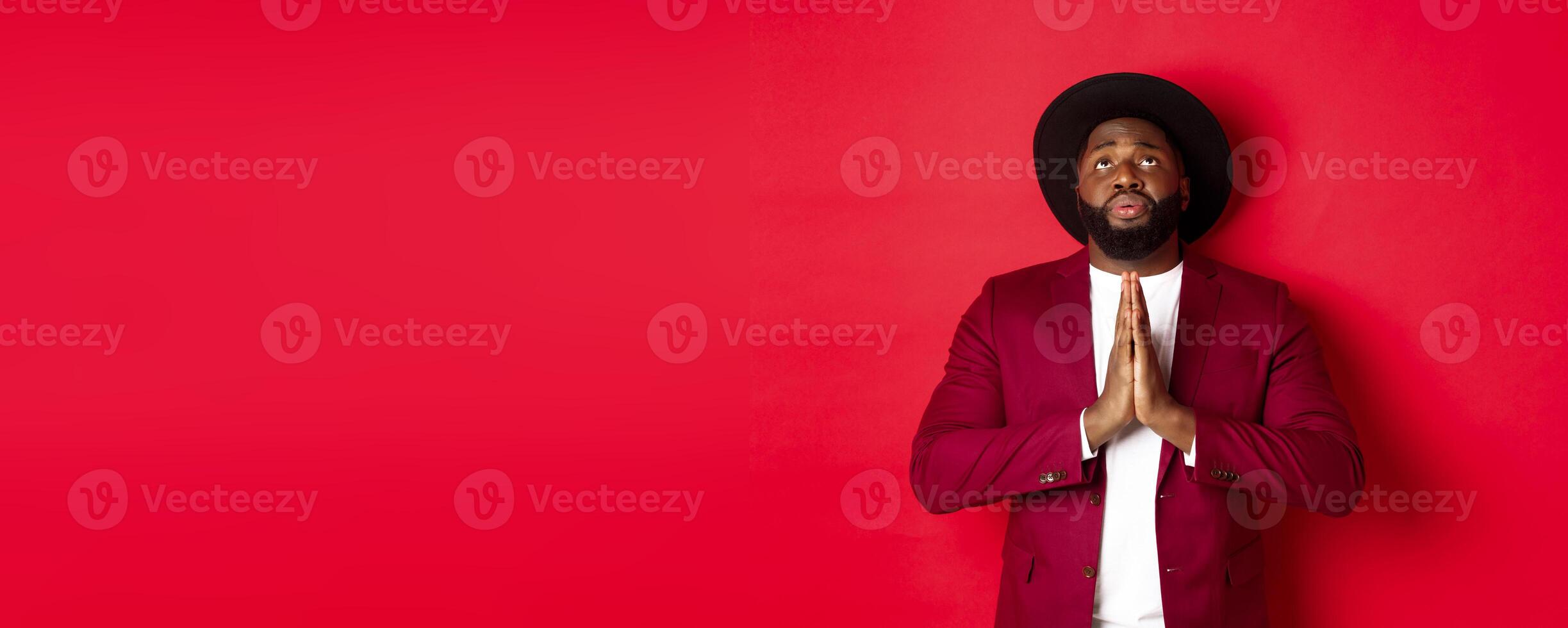 Hopeful and sad Black man talking to God, holding hands in pray and looking up, pleading, begging for help, standing against red background photo