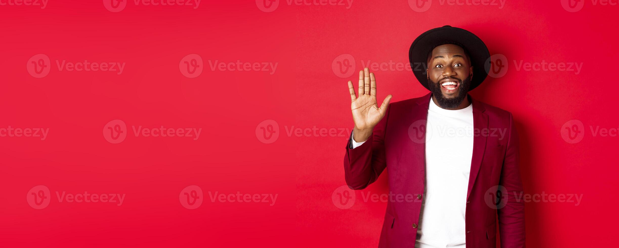 Friendly african american guy waving hand, saying hello and smiling, greeting you, standing over red background photo