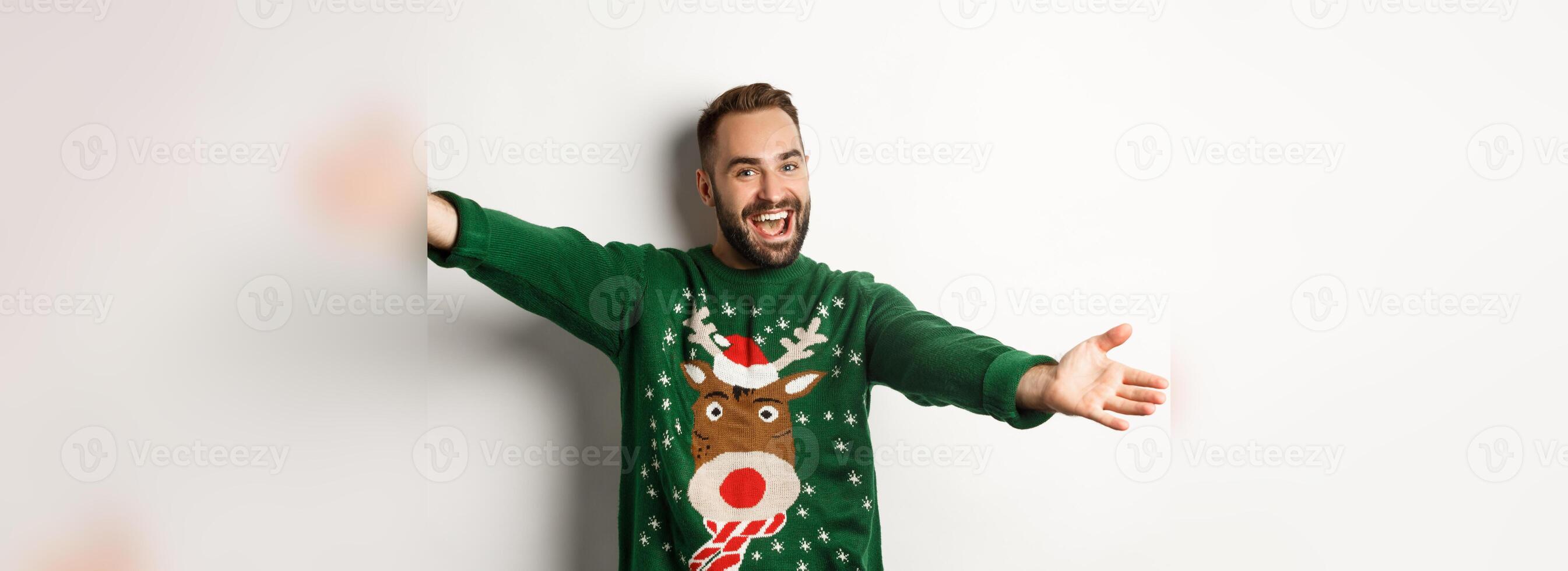 New year celebration and winter holidays concept. Friendly man spread hands for hug, welcome to Christmas party, wearing funny sweater, white background photo