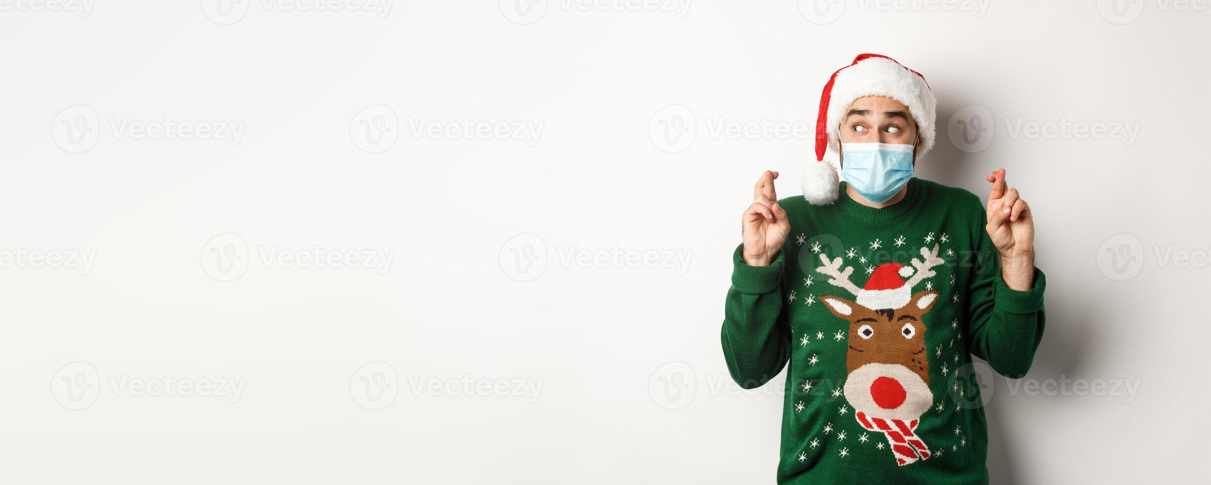 Concept of covid-19 and Christmas holidays. Excited man in face mask and Santa hat cross fingers, making a wish, standing over white background photo