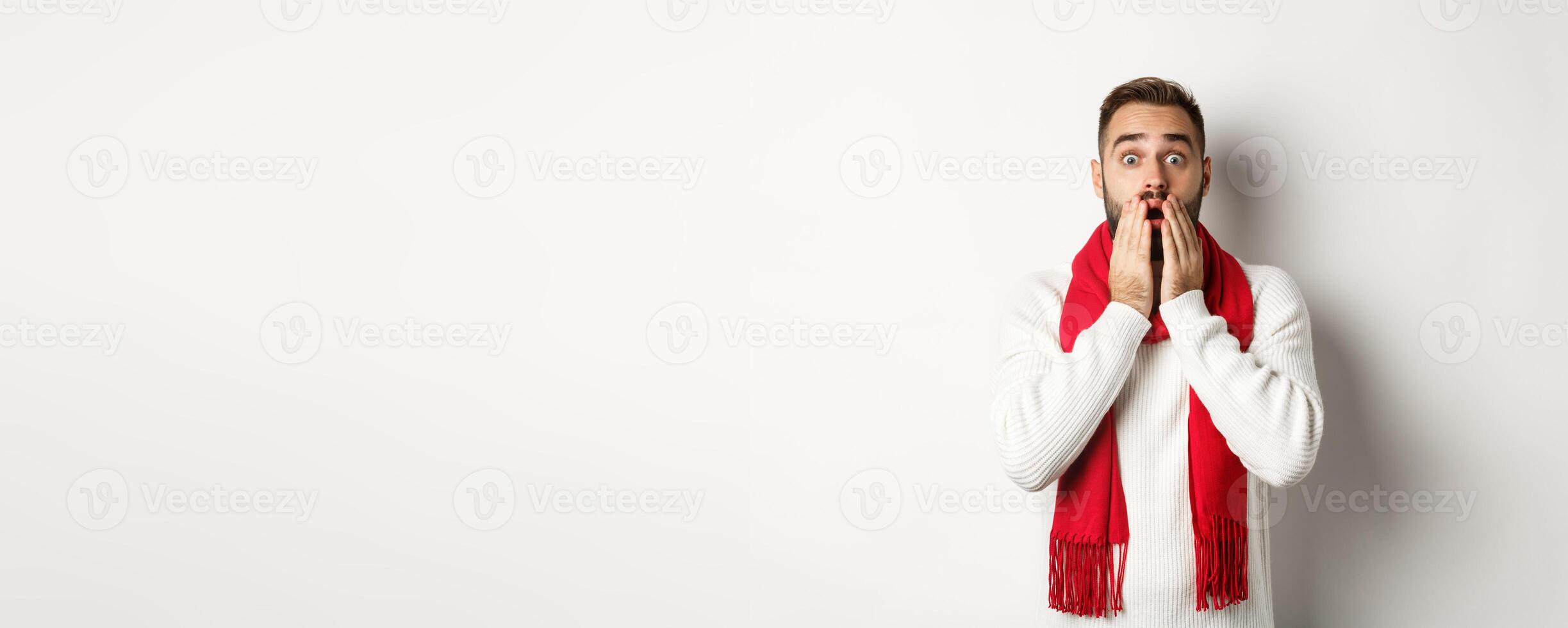 Christmas holidays and celebration concept. Handsome bearded guy in red scarf and sweater looking shocked, impressed with advertisement, white background photo