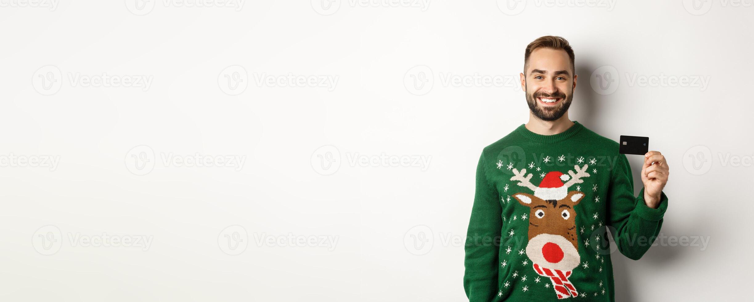 Christmas holidays and shopping concept. Young man in winter sweater showing credit card, standing over white background photo