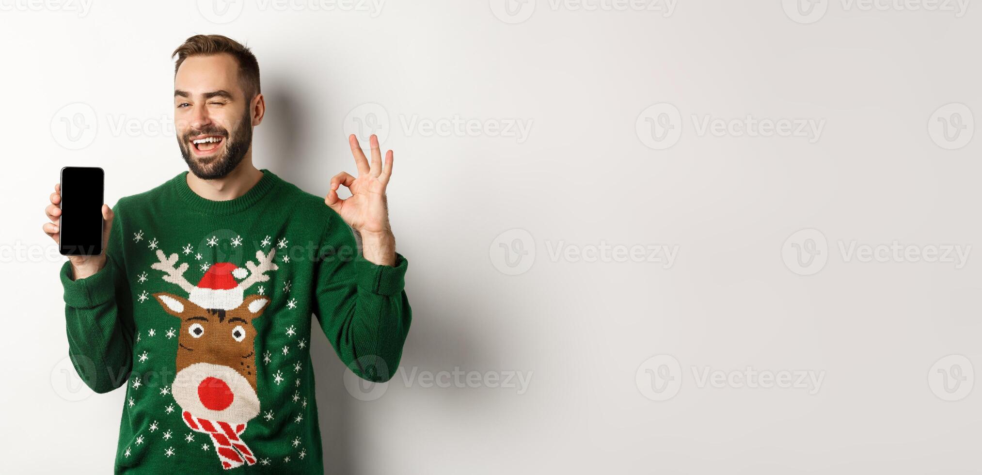 Christmas holidays and shopping concept. Confident guy winking and showing okay sign with mobile screen, standing over white background photo