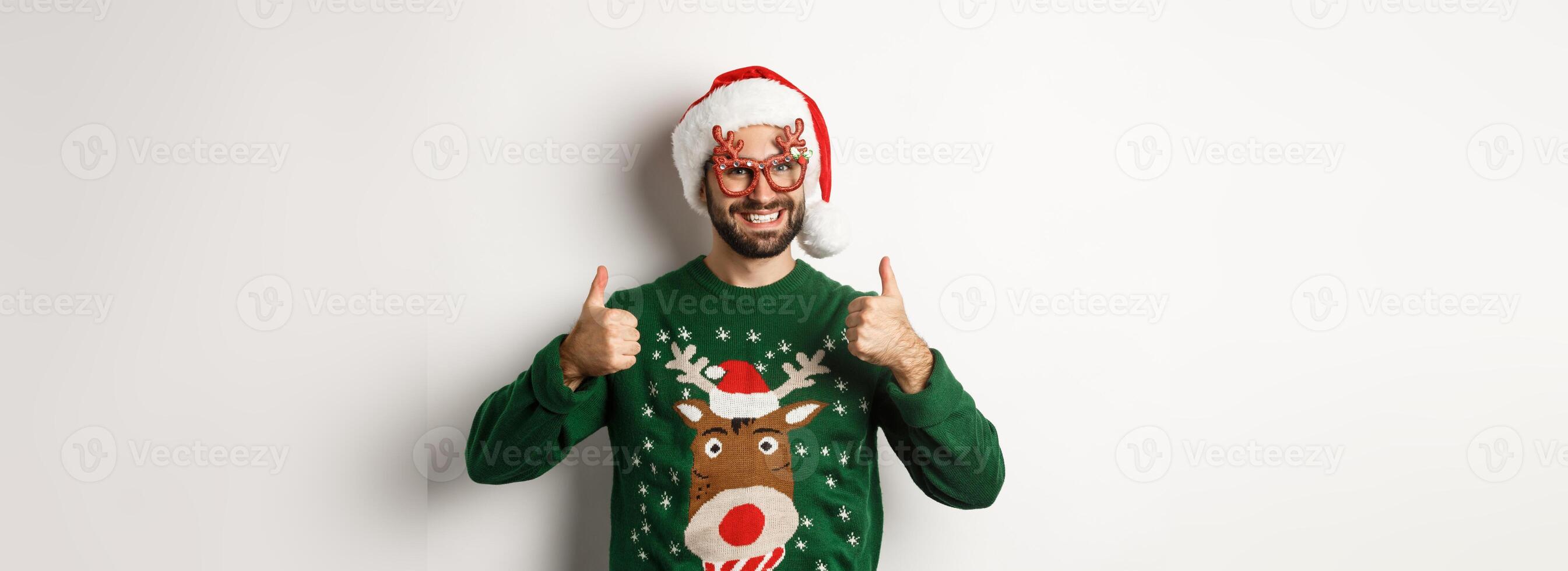 Christmas holidays, celebration concept. Happy man in Santa hat and party glasses showing thumbs up in approval, like something, standing over white background photo