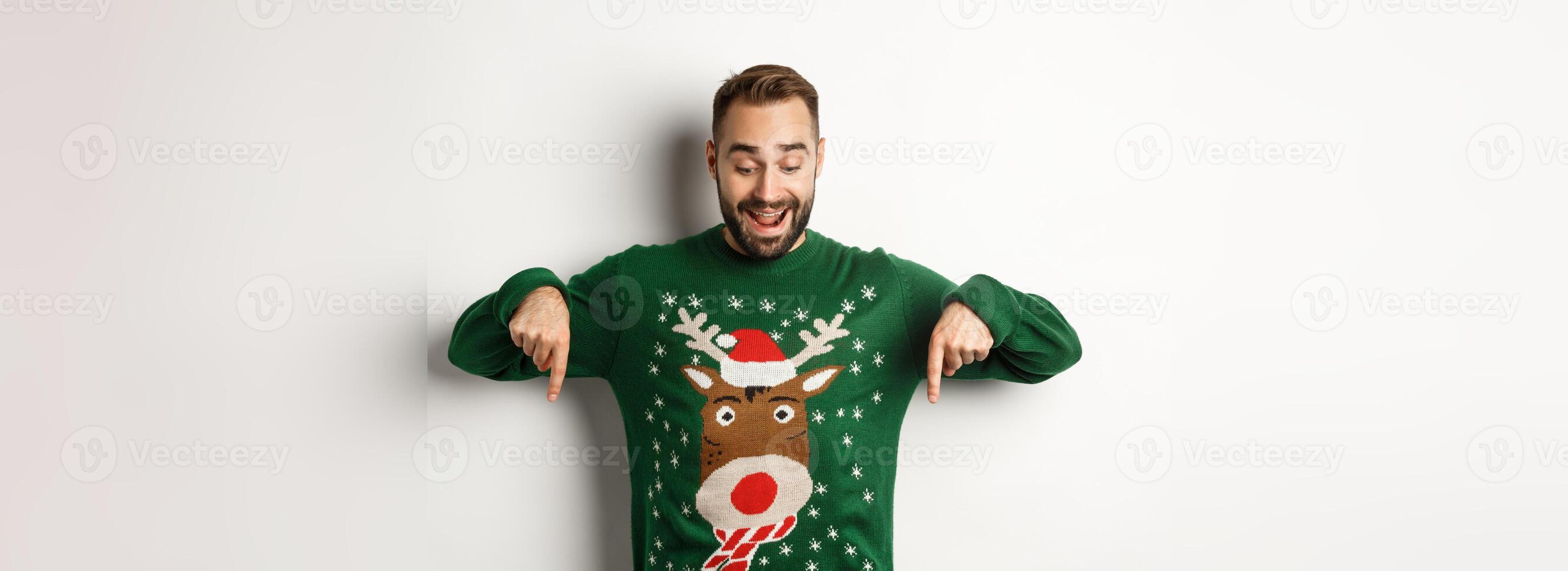 Winter holidays and christmas. Happy bearded man in green sweater, looking and pointing fingers down at advertisement, showing copy space, white background photo