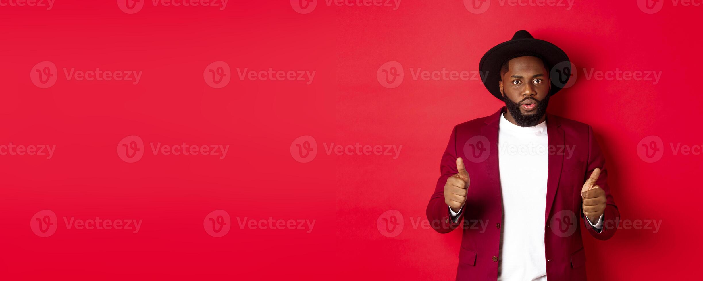 Impressed Black man showing thumbs up and staring amazed at camera, approve and praise something good, red background photo