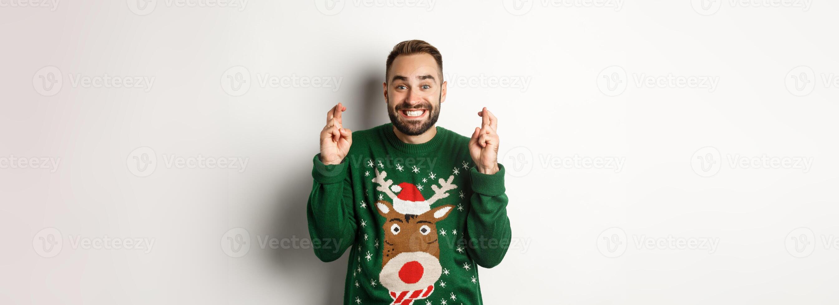 Christmas, holidays and celebration. Hopeful happy guy making wish, keep fingers crossed for good luck and smiling, standing over white background photo