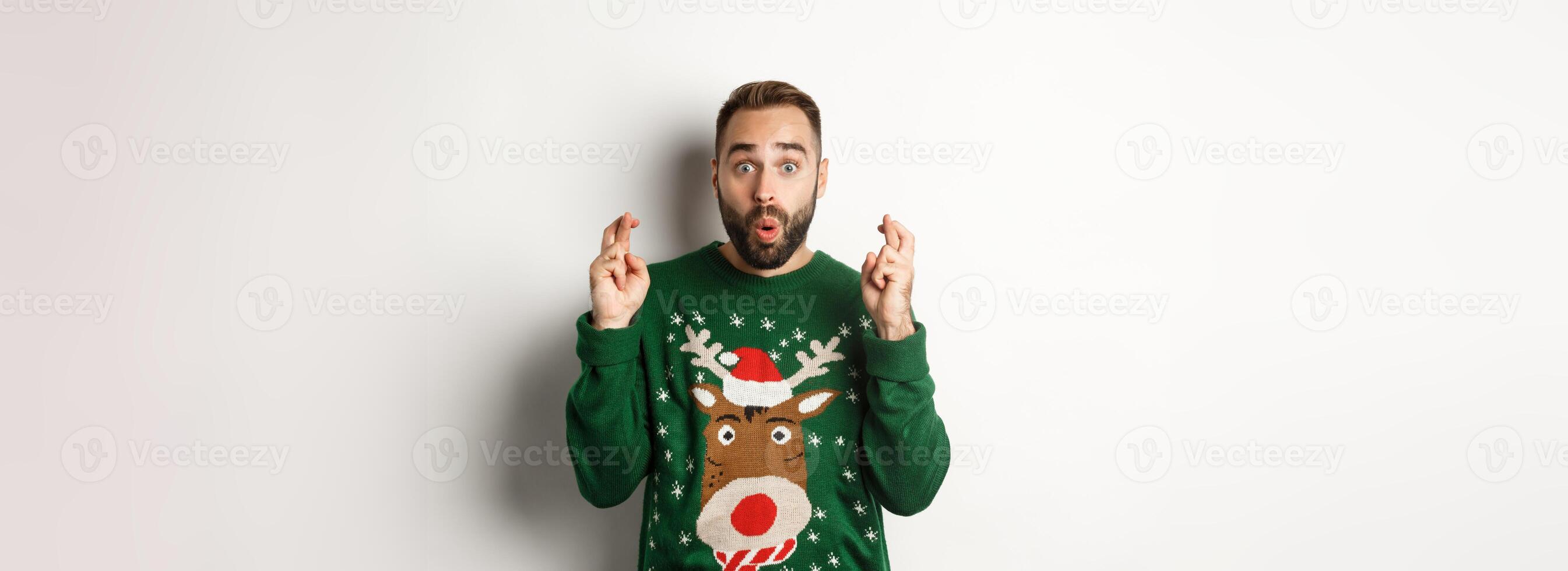 Christmas, holidays and celebration. Excited guy making wish, keep fingers crossed for good luck and smiling, standing over white background photo