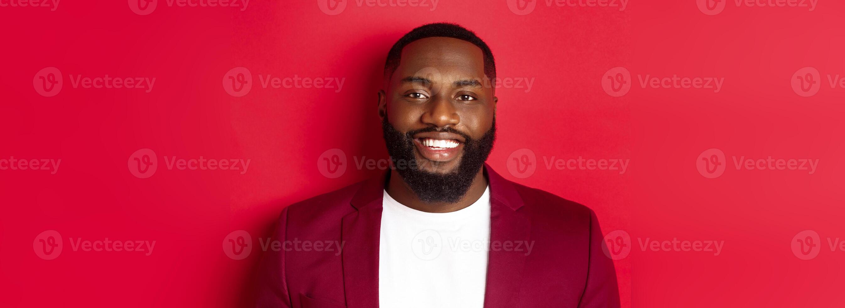 Close-up of handsome and stylish Black man wearing party blazer, smiling happy at camera, standing against red background photo