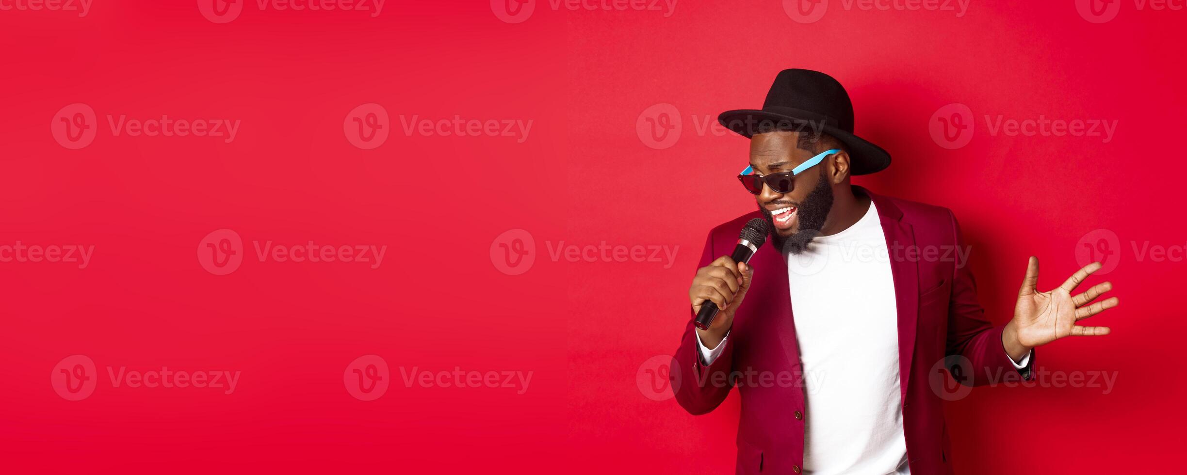 Passionate black male singer performing against red background, singing into microphone, wearing party outfit, standing over red background photo