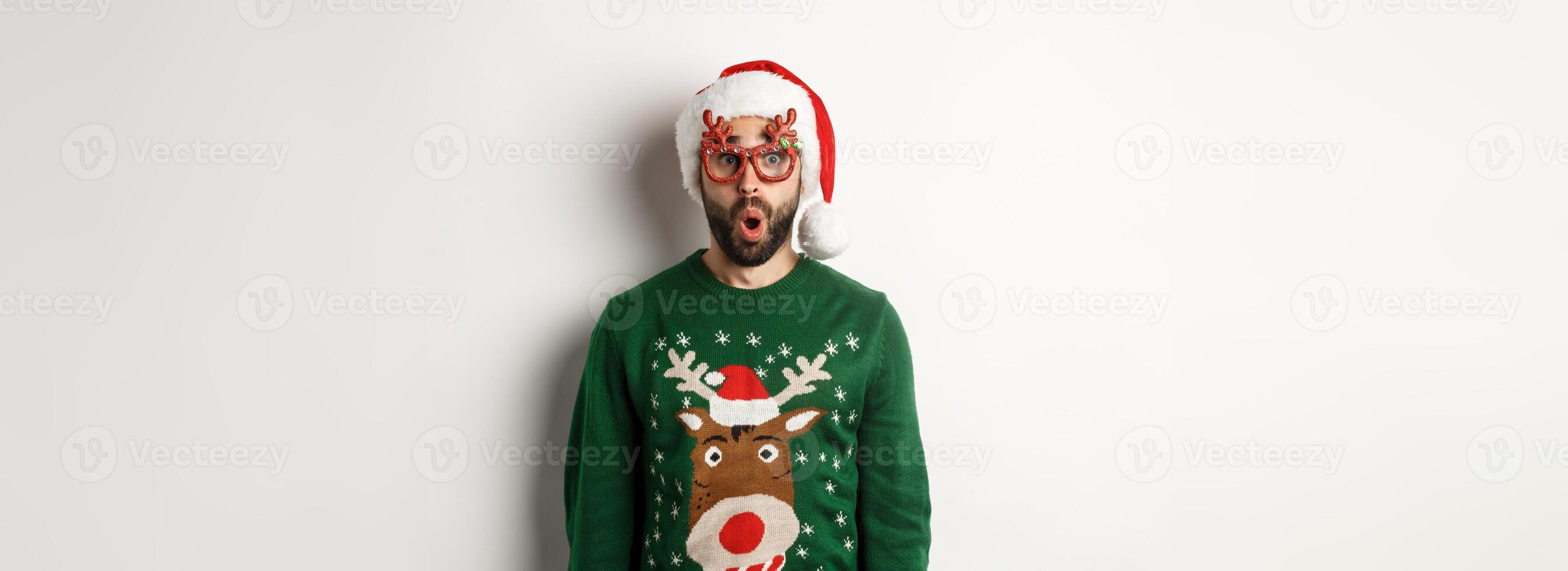 Christmas holidays, celebration concept. Surprised man in Santa hat and party glasses looking at something awesome, standing over white background photo